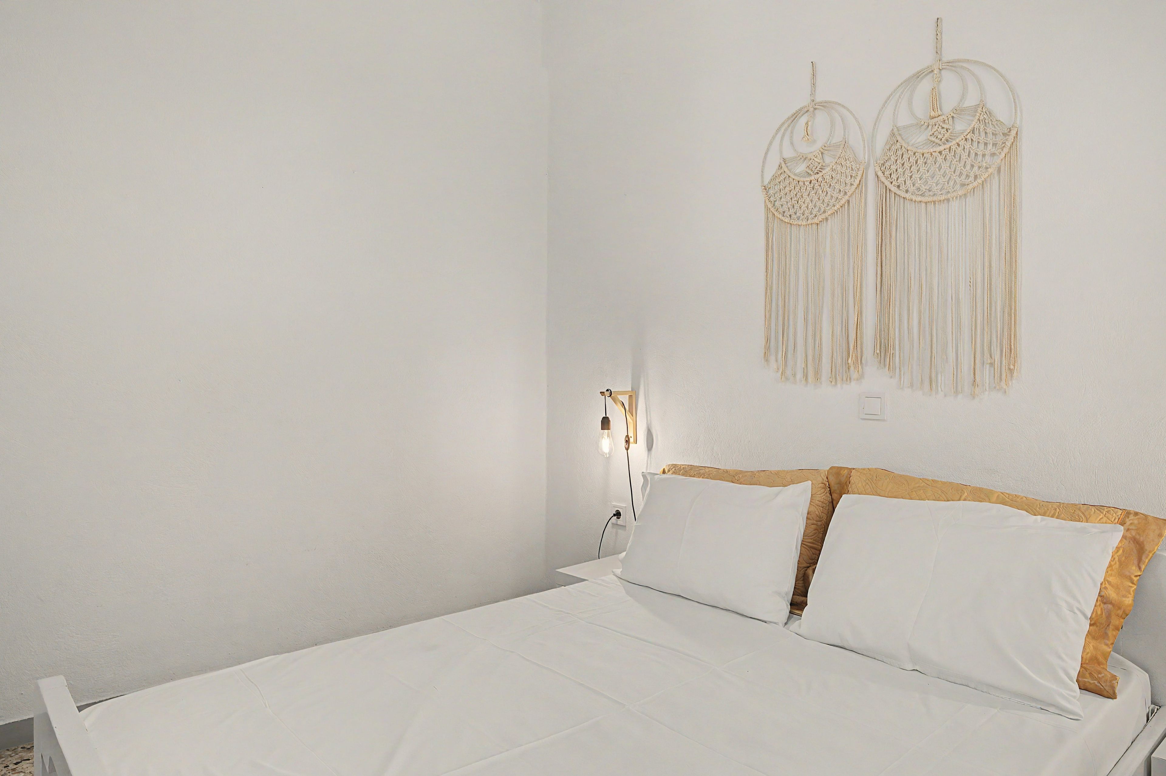 A bed with white bedding and wooden headboard, a wall-mounted light fixture, and macramé wall art.