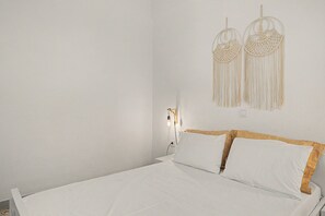 Una cama con ropa blanca y cabecera de madera, un aplique de pared y una decoración de macramé en la pared.