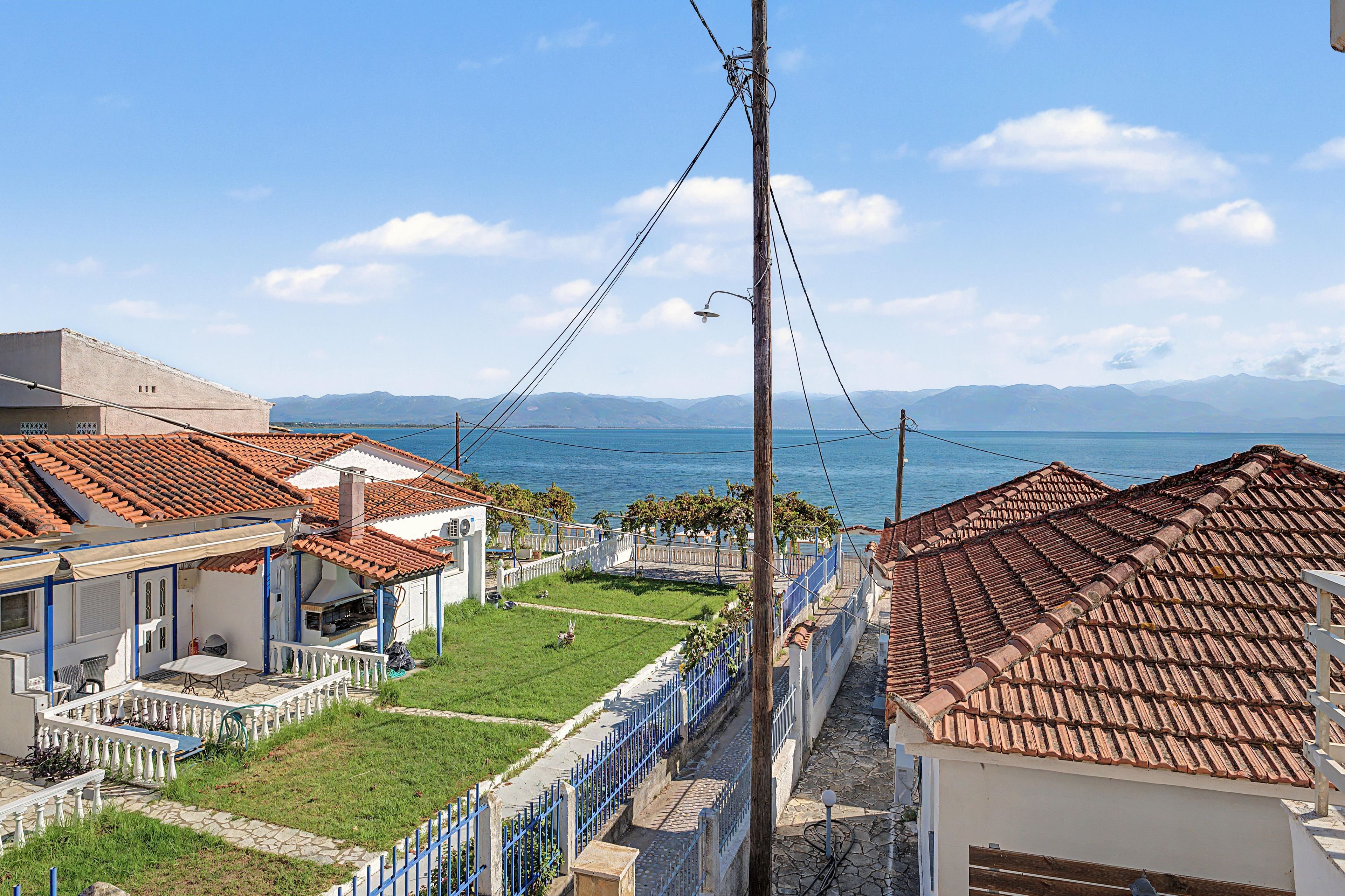 A coastal residential area with terracotta-tiled roofs, a grassy lawn, and a view of the sea and distant mountains.