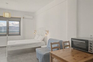A bedroom with a bed, a chair, a wooden table, and a kitchenette area with a microwave. - Apartment 'Kagias  2' with Sea View, Wi-Fi and Air Conditioning (Petalidi)