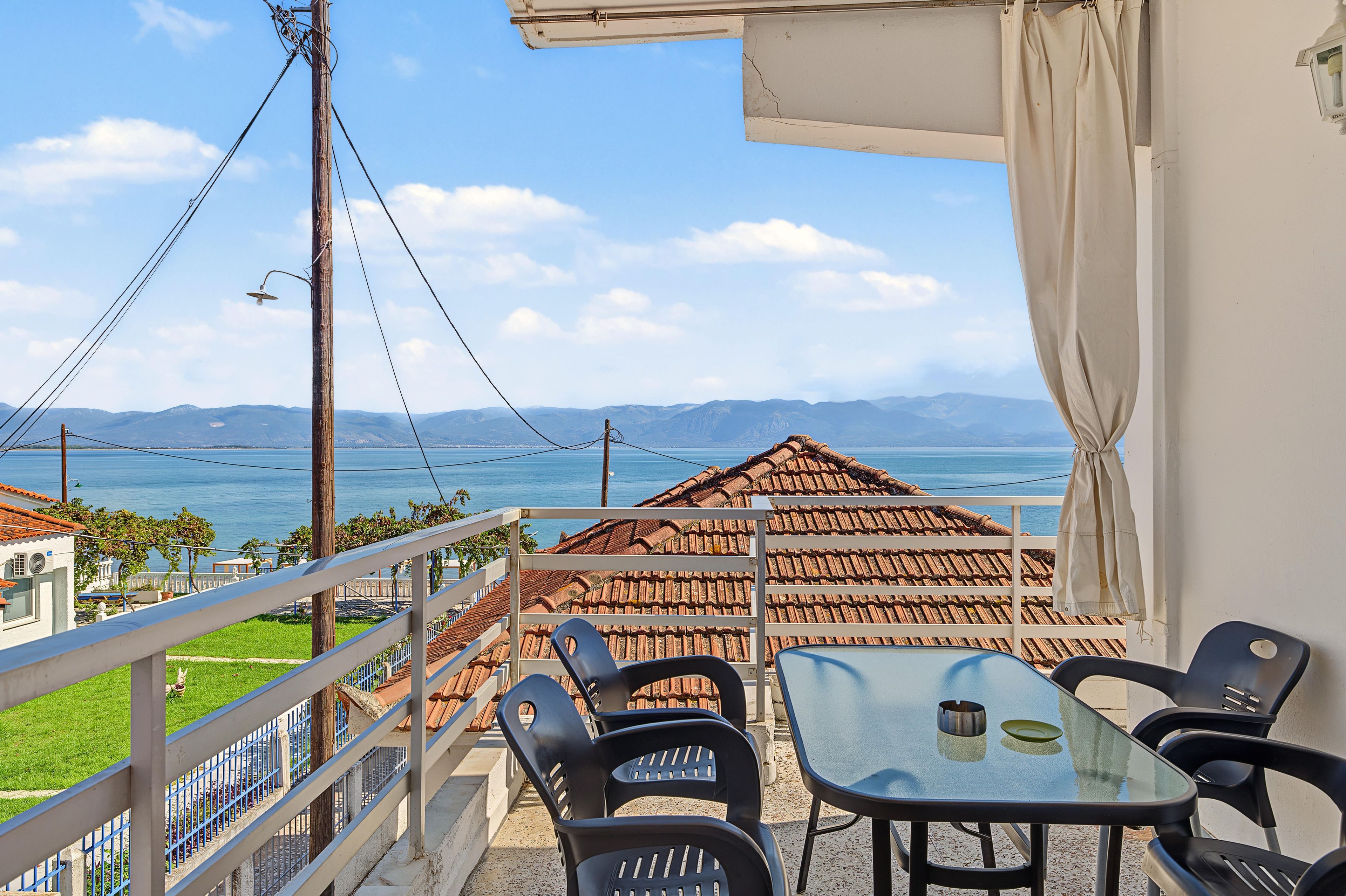 A balcony with a table and chairs, overlooking a coastal view with mountains in the distance.