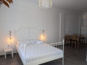 A white metal bed with a headboard, bedside lamps, a small white nightstand, and a wooden dining table with chairs. - Apartment 'Kagias  2' with Sea View, Wi-Fi and Air Conditioning (Petalidi)