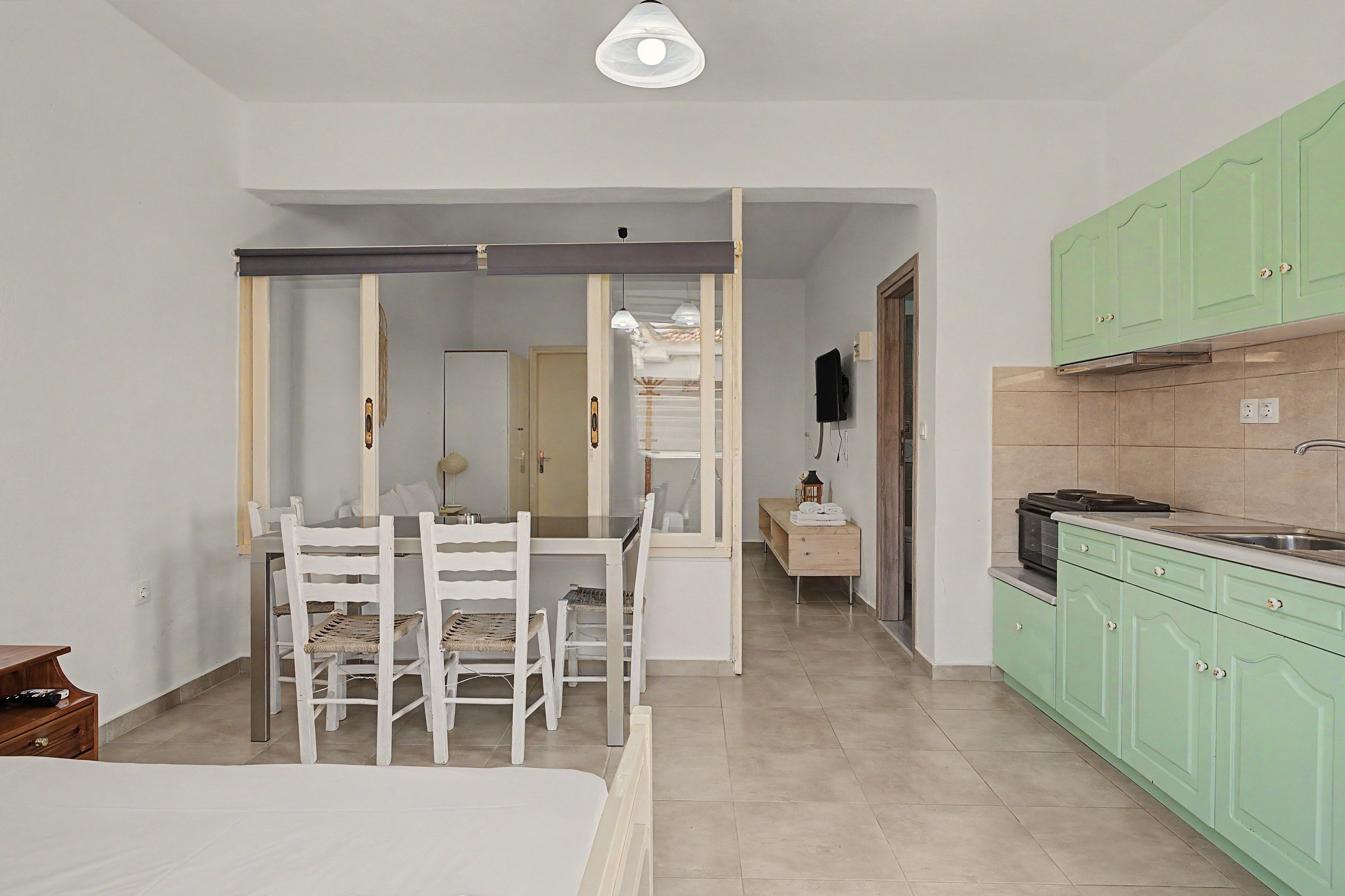 A room with a bed, a dining table with chairs, a kitchen with green cabinets, and a hallway leading to another room.