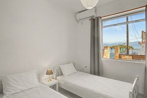 A bedroom with two beds, a window with a view of buildings and mountains, and a ceiling fan. - Apartment 'Kagias  4' with Sea View, Wi-Fi and Air Conditioning (Petalidi)