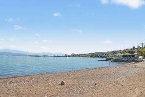 Ein kiesiger Strand mit ruhiger See, ein kleiner Felsen und eine in der Ferne liegende Stadt mit Gebäuden und Bergen.