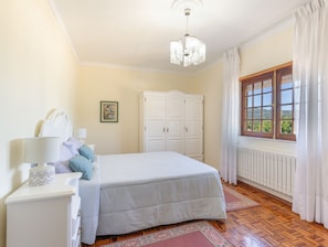 A bedroom with a bed, a dresser, a nightstand, a chandelier, a wardrobe, a window with curtains, and a radiator.