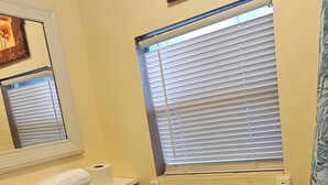 Un baño con inodoro, un lavabo con toalla, una ventana con persianas y un cuadro enmarcado sobre la ventana.