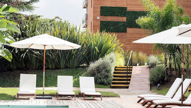 Zona junto a la piscina con sillones de descanso y sombrillas, rodeada de exuberante vegetación y una terraza de madera.