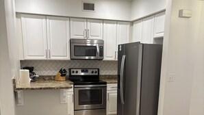 A kitchen with white cabinets, stainless steel appliances, a trash can, and a dining table with plates. - Enjoy beach life in a 3-bedroom condo in gorgeous Blue Mountain Beach on 30a. (Santa Rosa Beach)