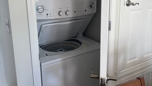 A compact, white washer and dryer unit with a control panel, a door, and a wooden chair in front. - Enjoy beach life in a 3-bedroom condo in gorgeous Blue Mountain Beach on 30a. (Santa Rosa Beach)