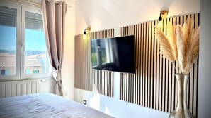 A bedroom with a bed, a TV mounted on a wooden panel wall, a window with curtains, and a vase with pampas grass. - Le Jardin Bleu, 6-person villa swimming pool (Saint-Geniès-de-Malgoirès)