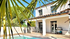 A poolside area with a clear blue pool, a covered patio with chairs, and a grill. - Le Jardin Bleu, 6-person villa swimming pool (Saint-Geniès-de-Malgoirès)