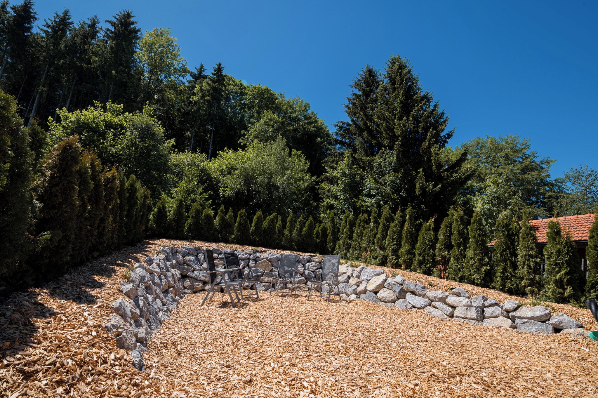 A landscaped area with a stone wall, outdoor seating, and a backdrop of tall trees.