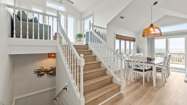 A spacious interior with a wooden staircase, white railings, a dining area with a round table, and large windows offering a view. - Walk to Beach | Sunset Views! Pool | Hot Tub| Duck (Duck)