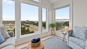 A spacious room with large windows, a grey sofa, a patterned rug, and a wicker side table. - Walk to Beach | Sunset Views! Pool | Hot Tub| Duck (Duck)