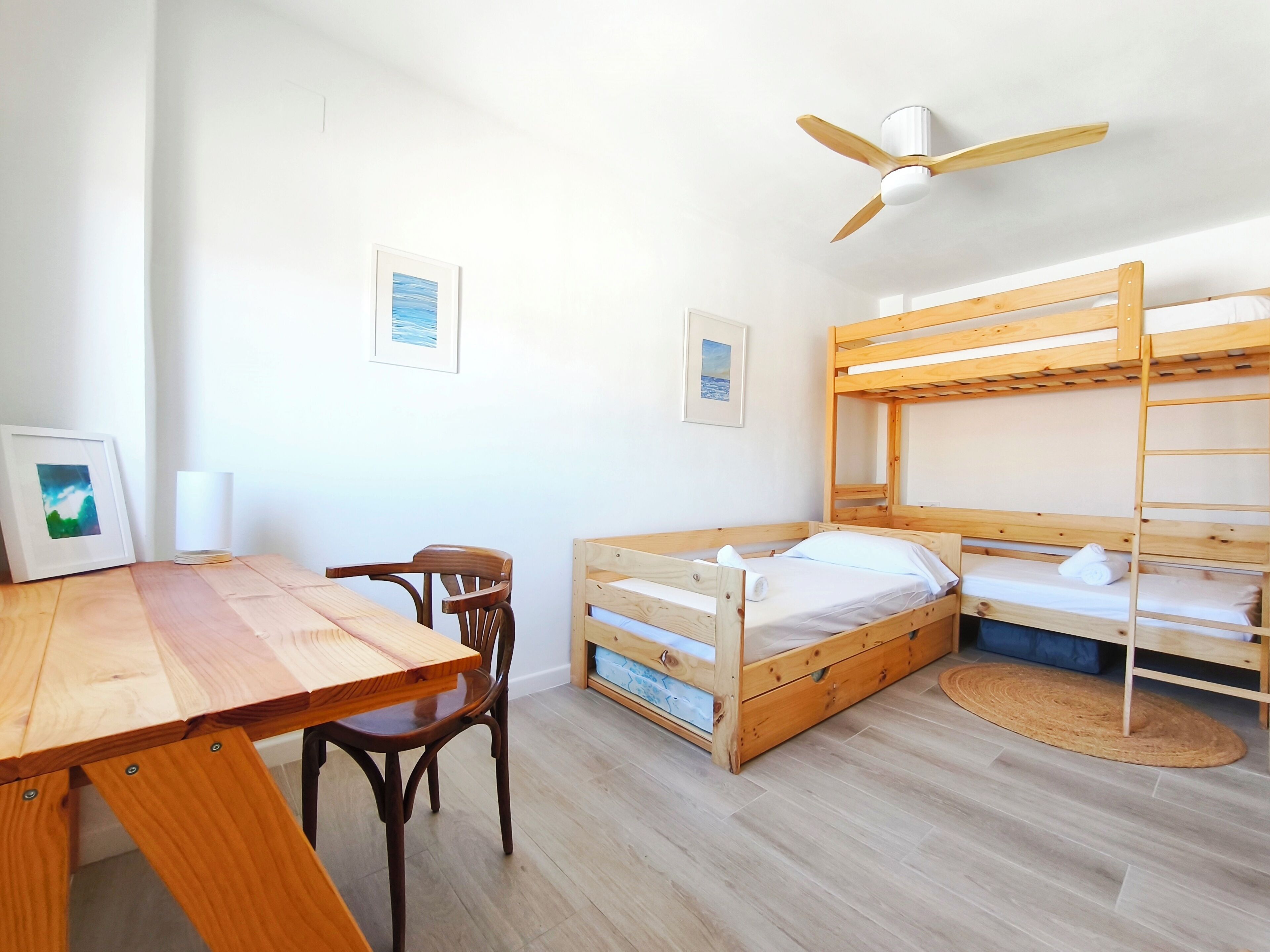A room with bunk beds, a wooden table, a chair, a ceiling fan, and framed pictures on the wall.