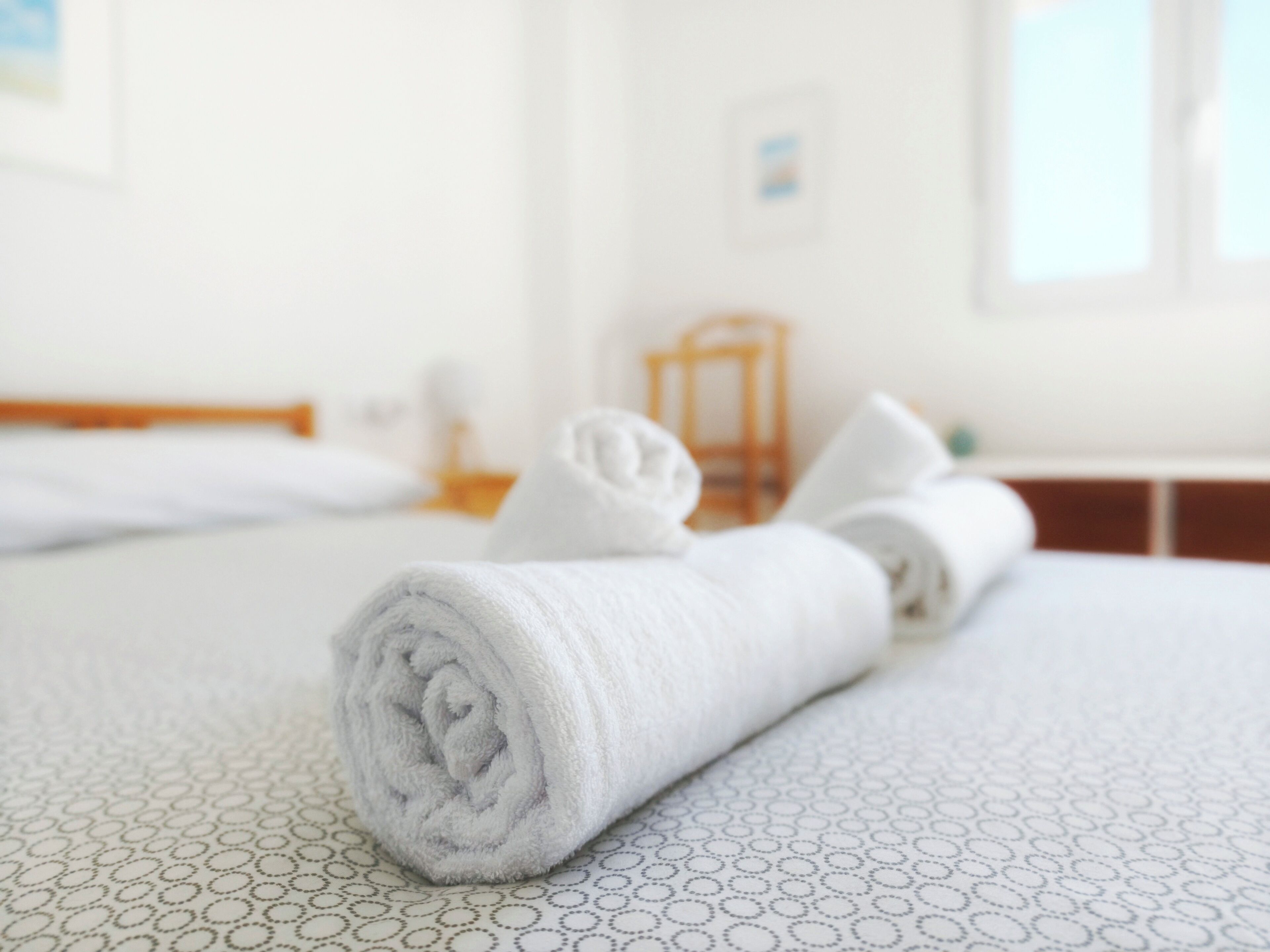A neatly made bed with rolled towels, a wooden headboard, and a small wooden chair in a bright room.