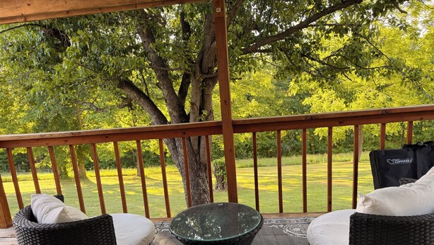 A covered outdoor seating area with wicker furniture and a glass table, overlooking a grassy landscape.