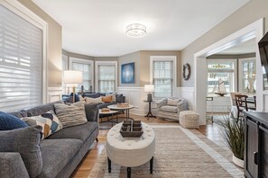 A living room with a sofa, ottoman, and lamps. There are windows with blinds, a chandelier, and a painting on the wall. - Historic Luxury Meets Modern Living — 6BR Victorian in the Heart of Newport (Newport)