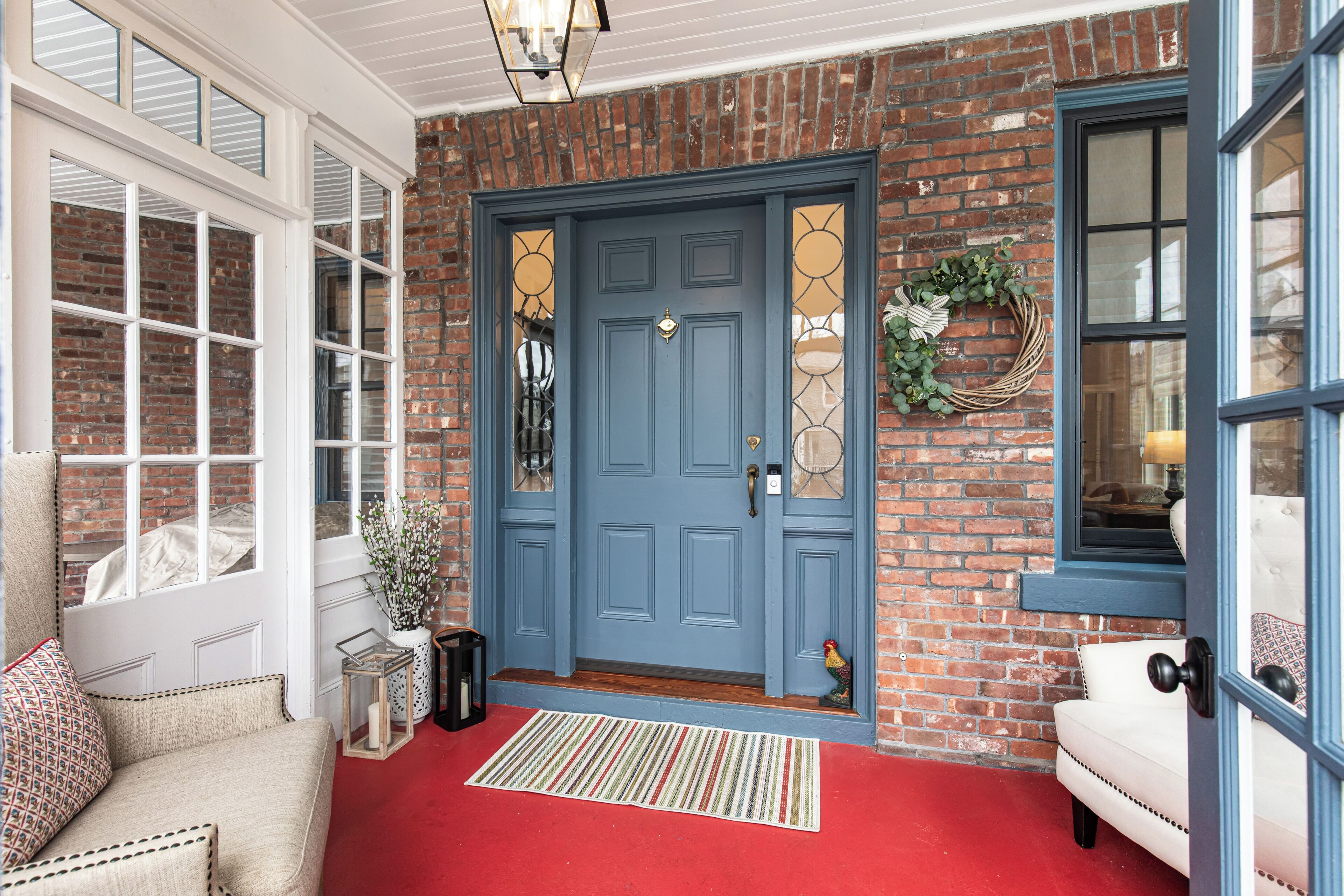 Fachada de ladrillo con puerta azul, ventanas enmarcadas en blanco, alfombra roja y banco blanco.