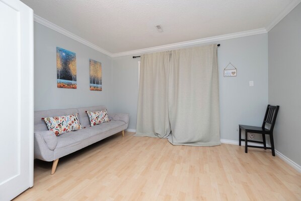 A living room with a grey sofa, two cushions, a wooden chair, a beige curtain, and two wall art pieces.