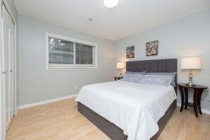 A bedroom with a bed, two bedside tables with lamps, a window with blinds, and two wall-mounted paintings.