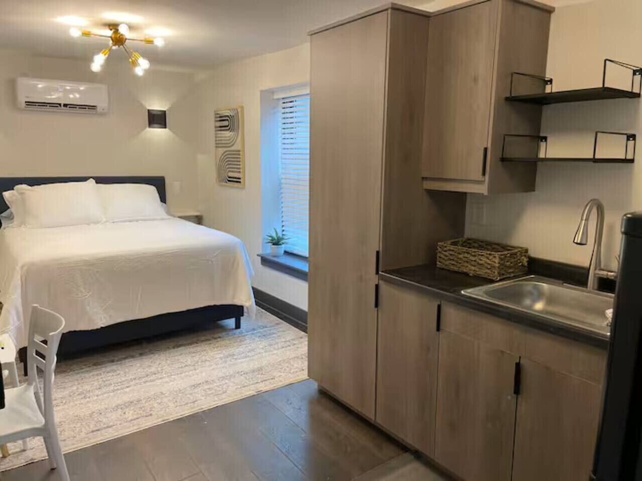 Ein Schlafzimmer mit Bett, einer kleinen Sitzecke, einer Küchenzeile mit Spüle und Schränken sowie einem Fenster mit Jalousien.