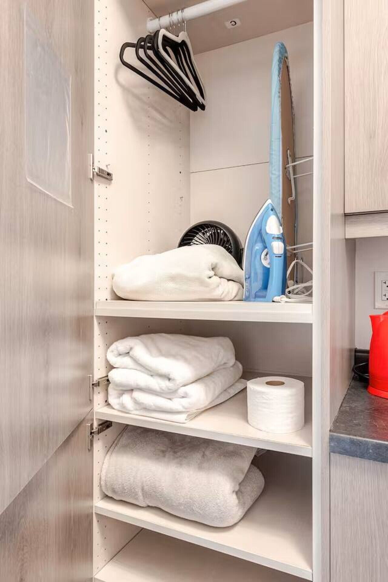 A closet with shelves holding folded towels, a blue iron, and a black fan.