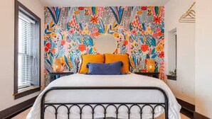 Un lit avec un cadre en métal, un mur décoré d’une fresque florale aux couleurs vives et une fenêtre avec des stores.
