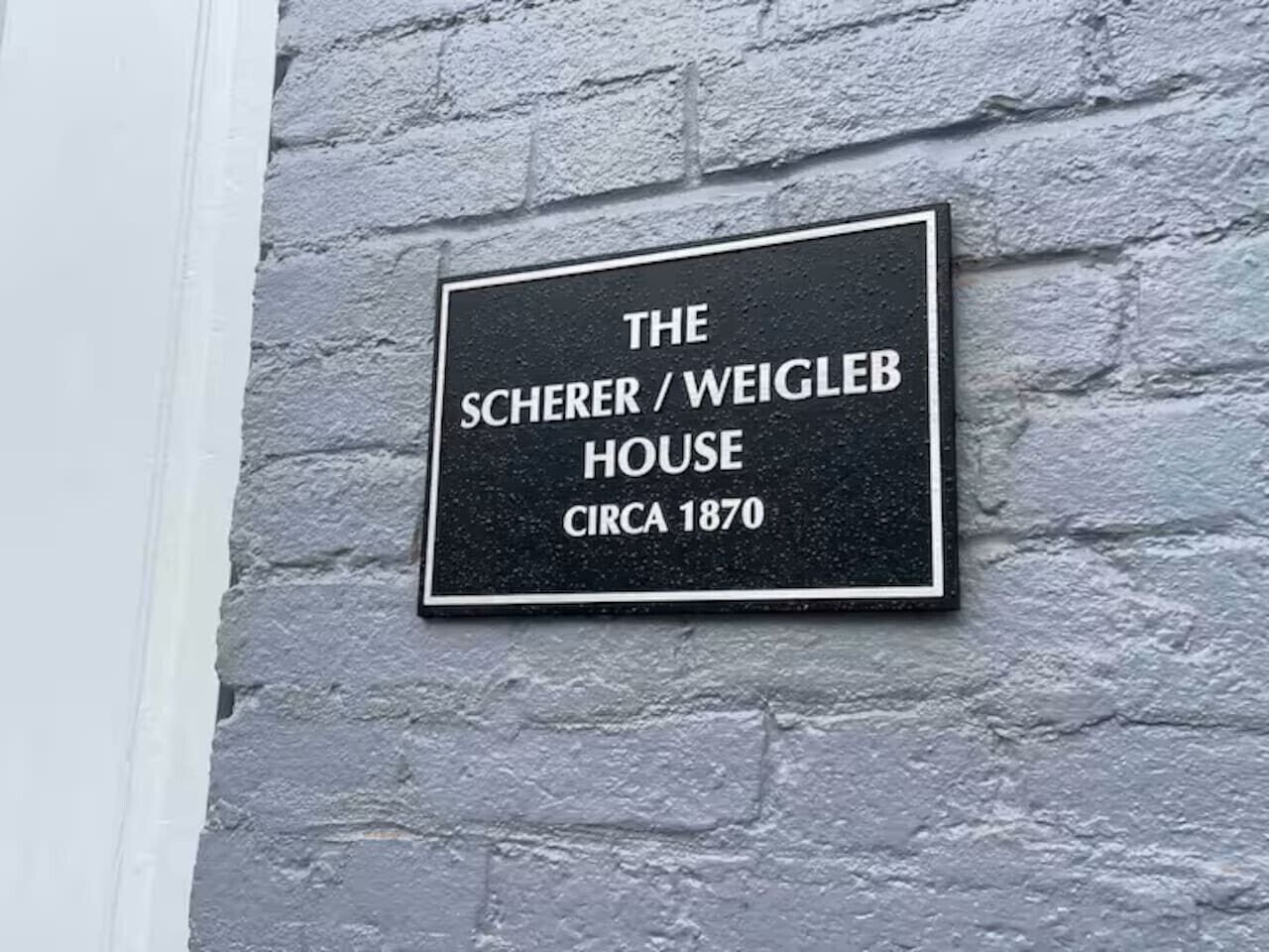 Une plaque fixée sur un mur à la texture particulière indique : « LA MAISON SCHERER / WEIGLEB, vers 1870 ».