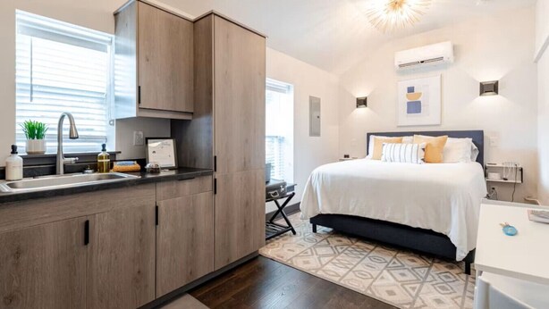Una habitación de hotel moderna con una zona de cocina, una cama con muchos cojines y una lámpara en el techo.