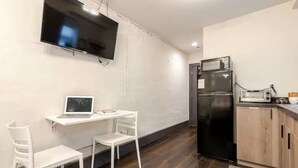 Una pequeña kitchenette equipada con refrigerador, microondas y tostadora. Hay un escritorio blanco con una laptop y dos sillas blancas. Un televisor de pantalla plana está montado en la pared.