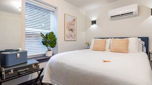 Un dormitorio con cama, ventana con persianas, planta en maceta, aire acondicionado de pared y maleta sobre un escritorio.