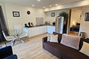A modern living room with a glass dining table, a sofa, a kitchen area with a refrigerator, and a small dining table.