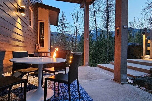 An outdoor patio with a round table and chairs, a fire feature, and a view of trees and mountains.