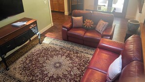 A living room with a leather sofa, a flat-screen TV, a patterned rug, and a kitchen visible through an archway.