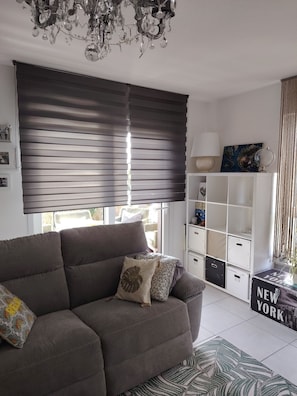 Ein Wohnzimmer mit einer Couch, einem Kronleuchter, einem Fenster mit Jalousien, einer Lampe und Stauraumeinheiten.