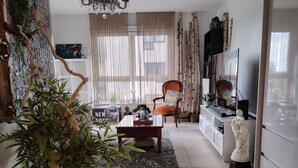Una sala de estar con sofá, mesa de centro y una butaca con cojín. Hay una planta en maceta, una araña y un televisor.