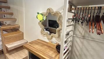 Une chambre avec un bureau en bois, un miroir, une télévision, une armoire avec des vêtements suspendus et un escalier.