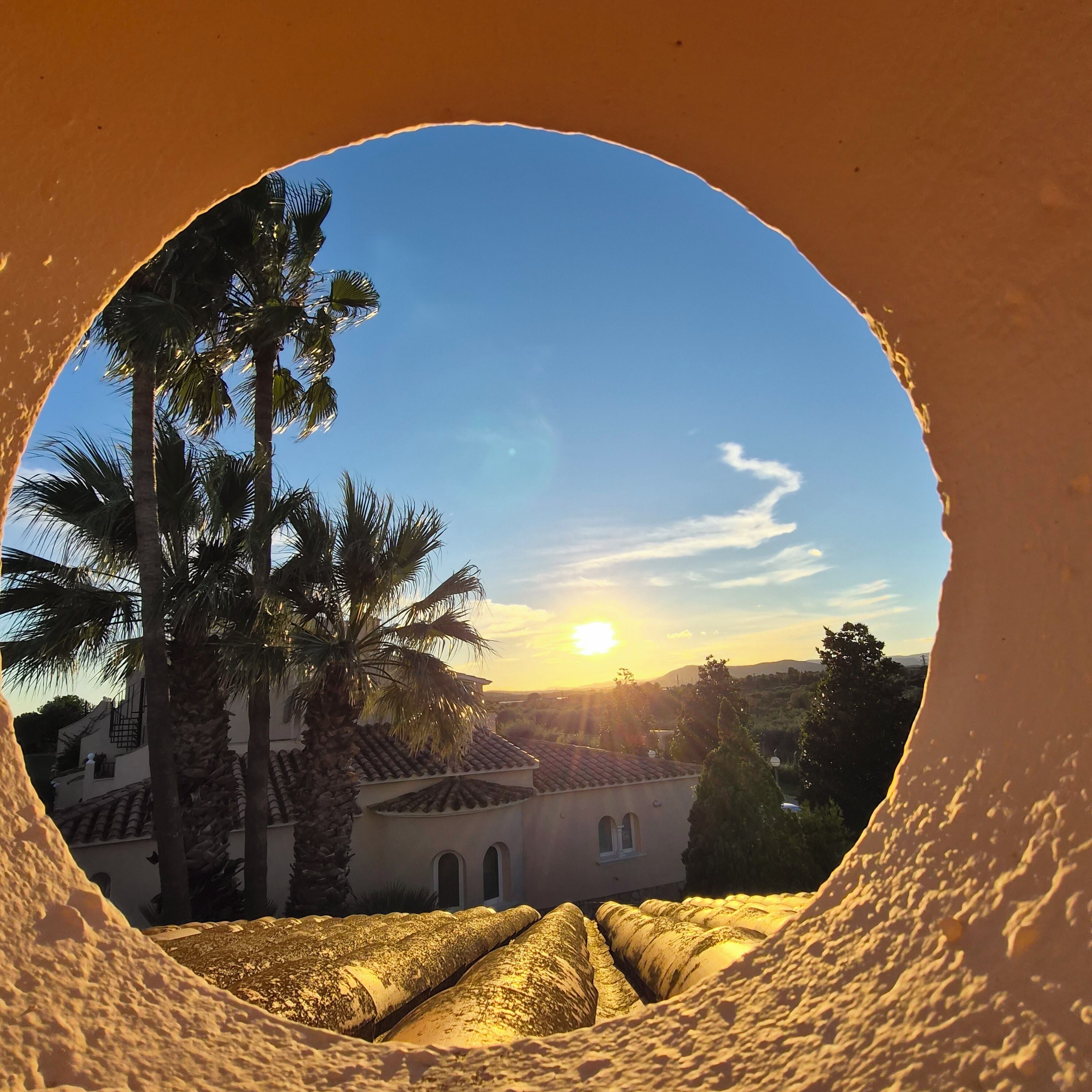 A circular view of a building with a tiled roof, palm trees, and a sunset.