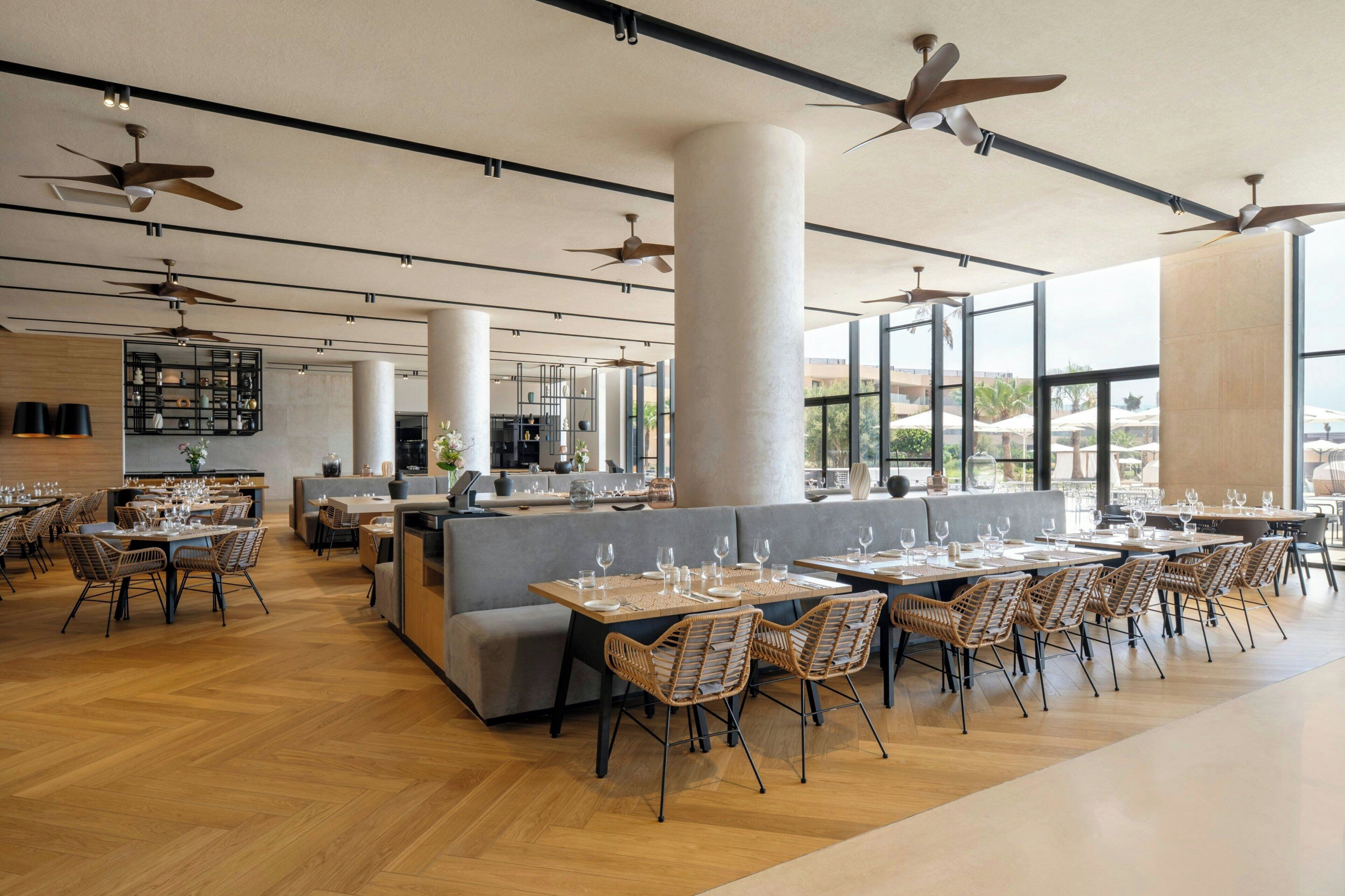 Un restaurante espacioso con techos altos, varios ventiladores de techo y grandes ventanales que ofrecen vistas al exterior.