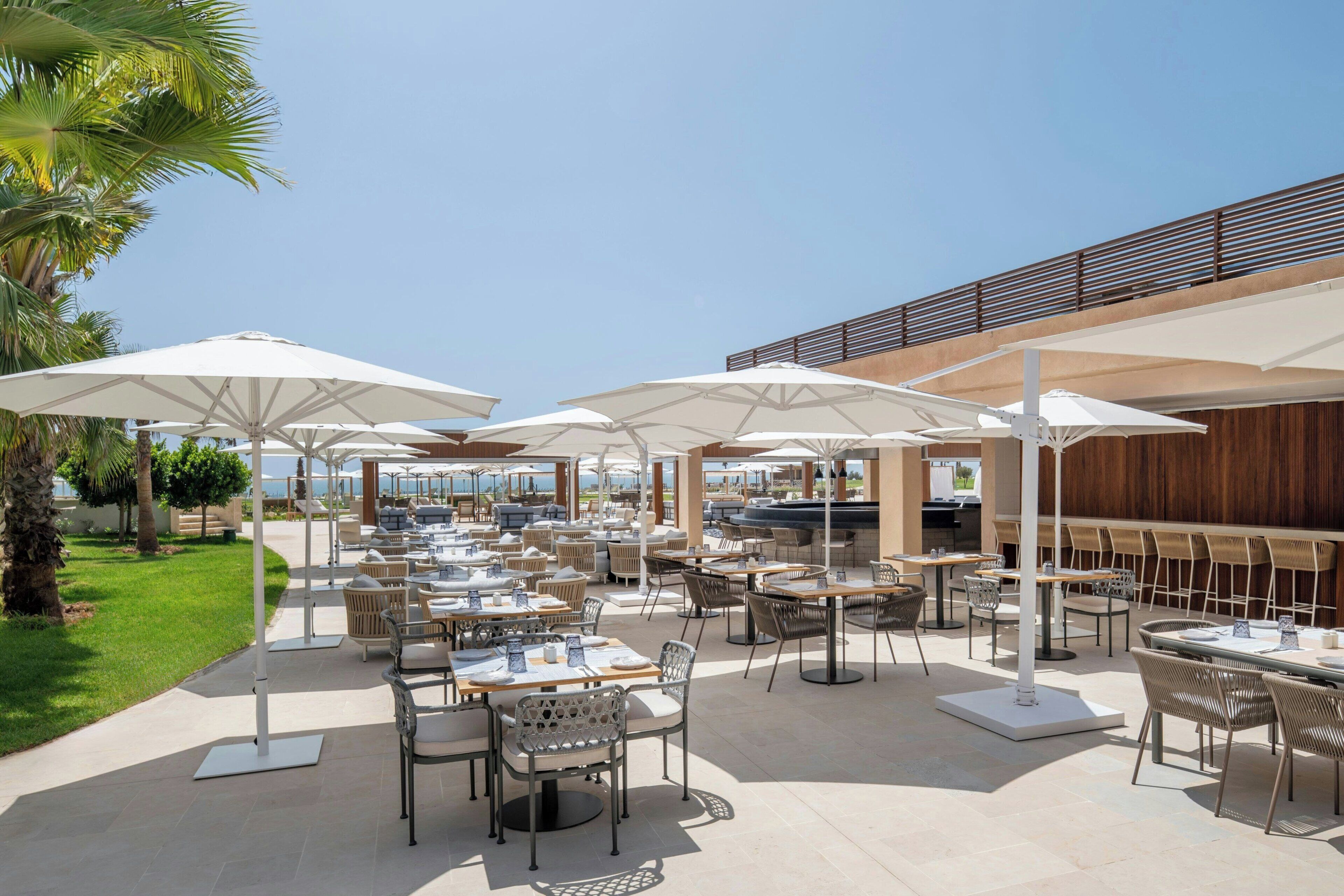 An outdoor dining area with multiple tables and chairs, umbrellas, and a view of the ocean.