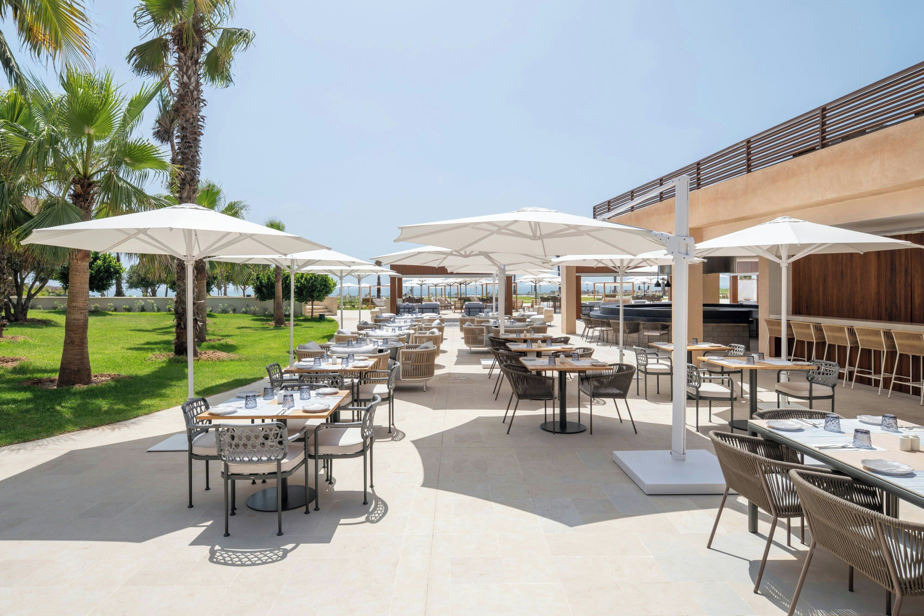 An outdoor dining area with tables and chairs, umbrellas, and palm trees.