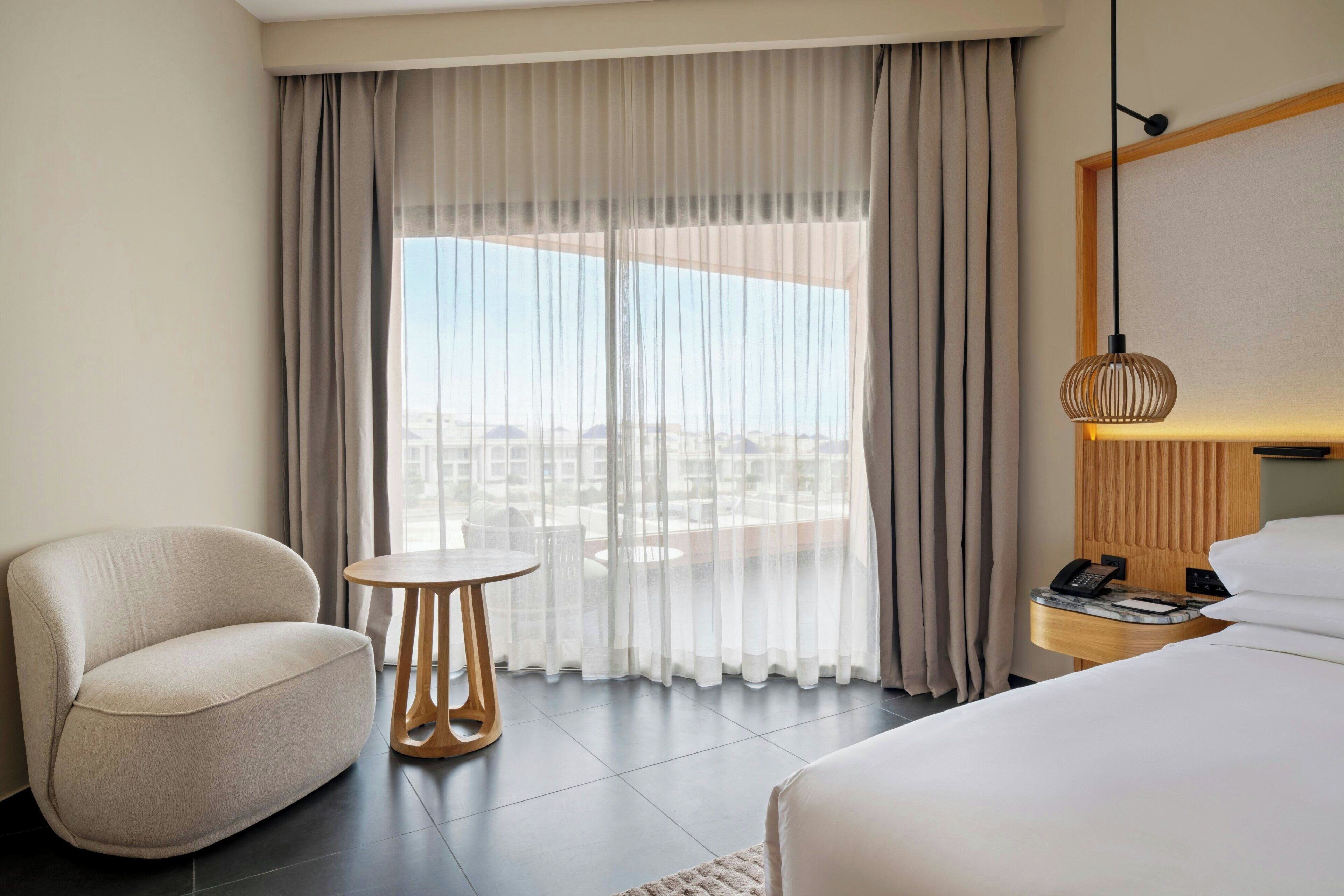Habitación de hotel con cama, una silla, una mesita redonda, lámpara y vistas a la ciudad a través de cortinas translúcidas.