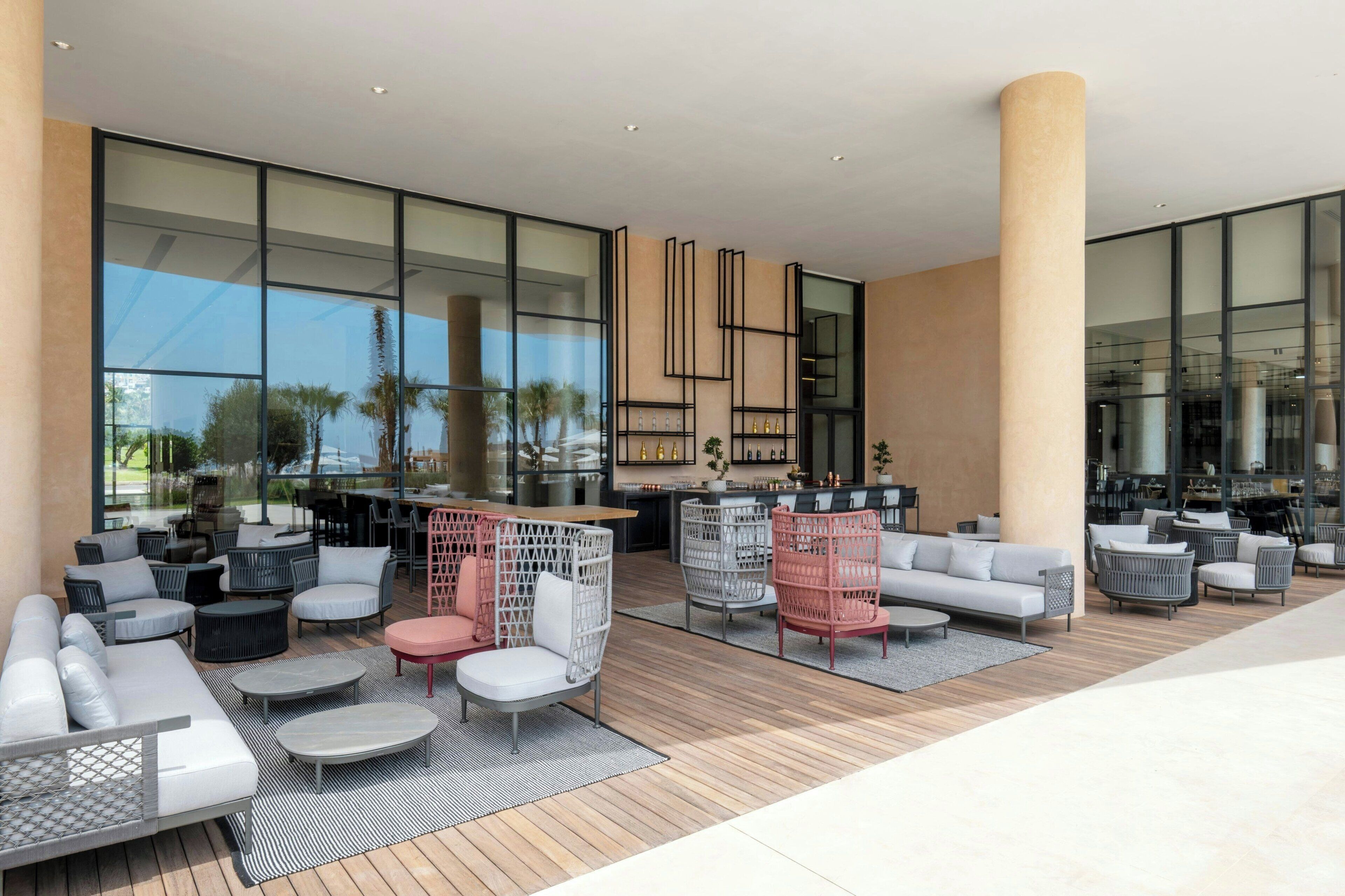A modern outdoor seating area with sofas, chairs, and tables, large windows, and a view of palm trees.