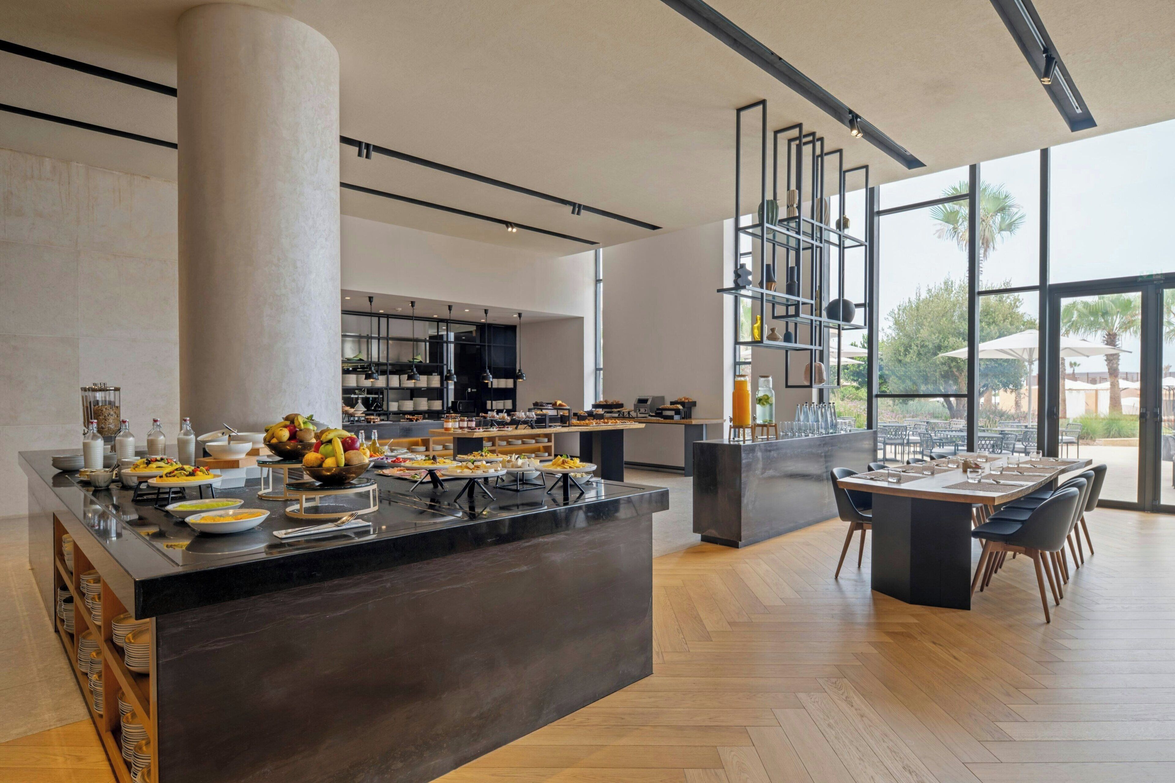 A modern kitchen with a central island, dining area with tables, and a view through large windows.