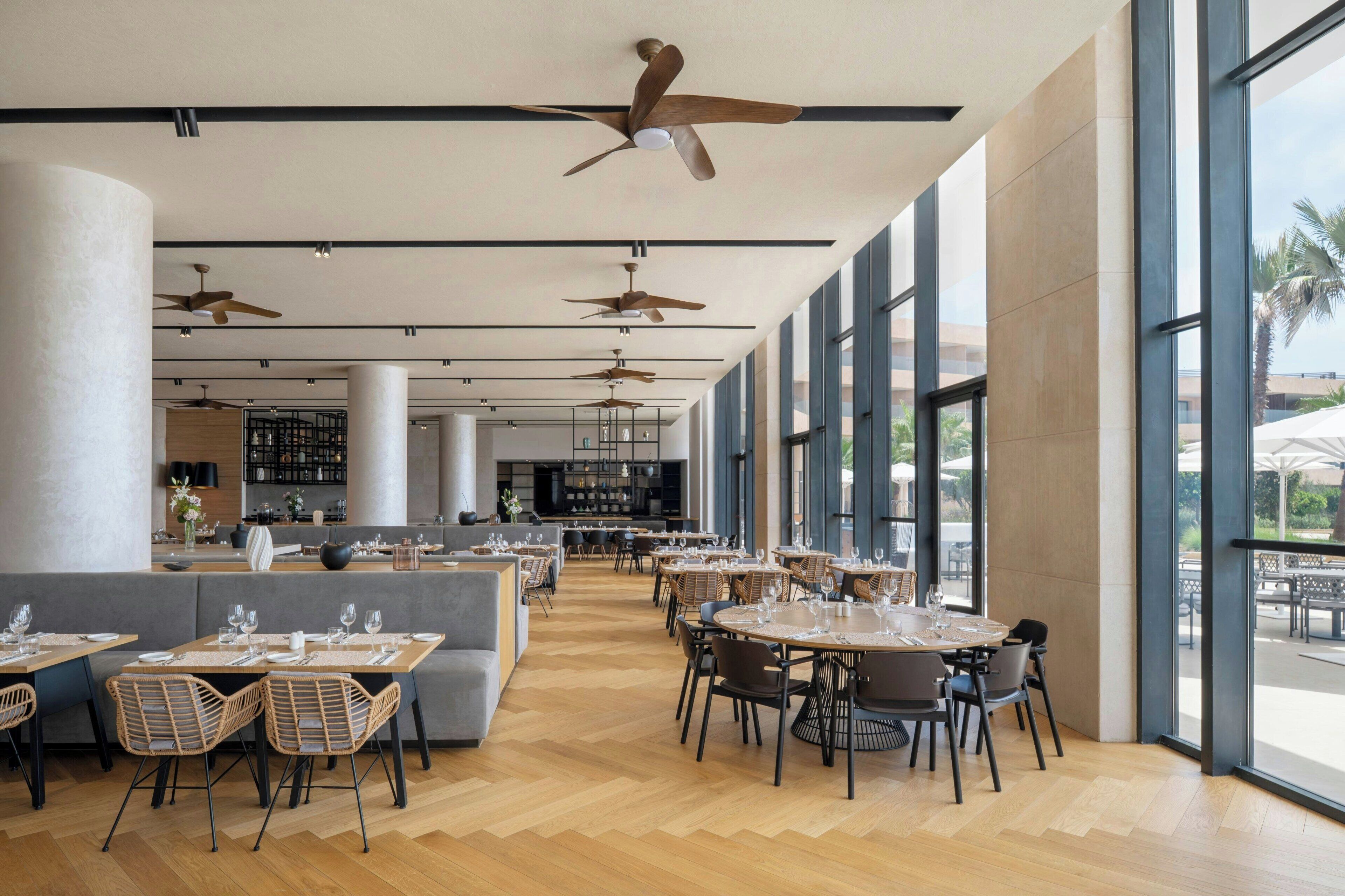 A spacious indoor dining area with multiple tables and chairs, ceiling fans, and large windows offering views of the outdoors.
