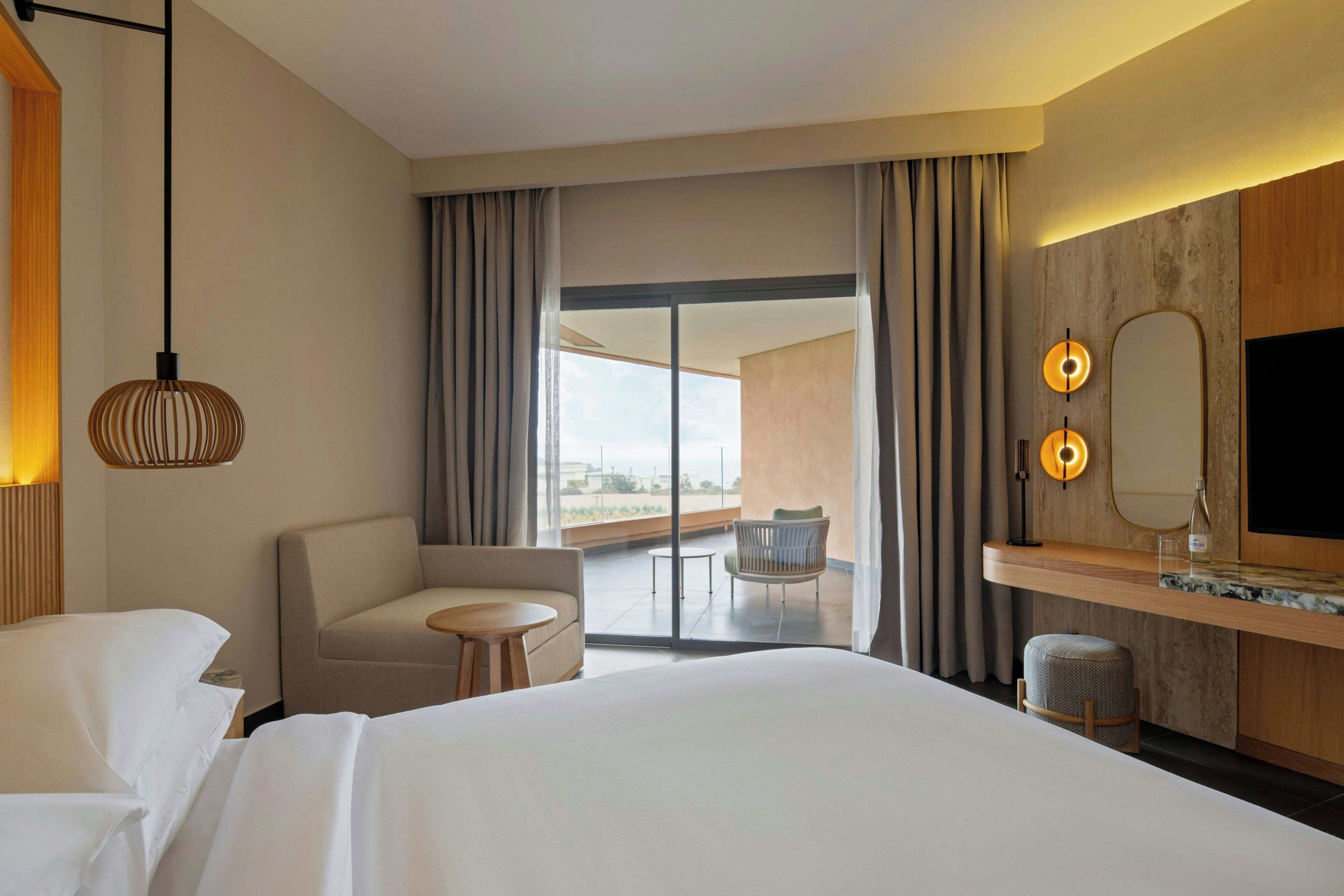 Une chambre d’hôtel dotée d’un grand lit, d’un coin salon avec un fauteuil et une petite table, d’un balcon avec vue et d’un téléviseur fixé au mur.