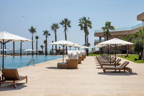 A poolside area with lounge chairs and umbrellas, palm trees, and a clear view of the ocean. - Taghazout Bay Marriott Resort (Taghazout)