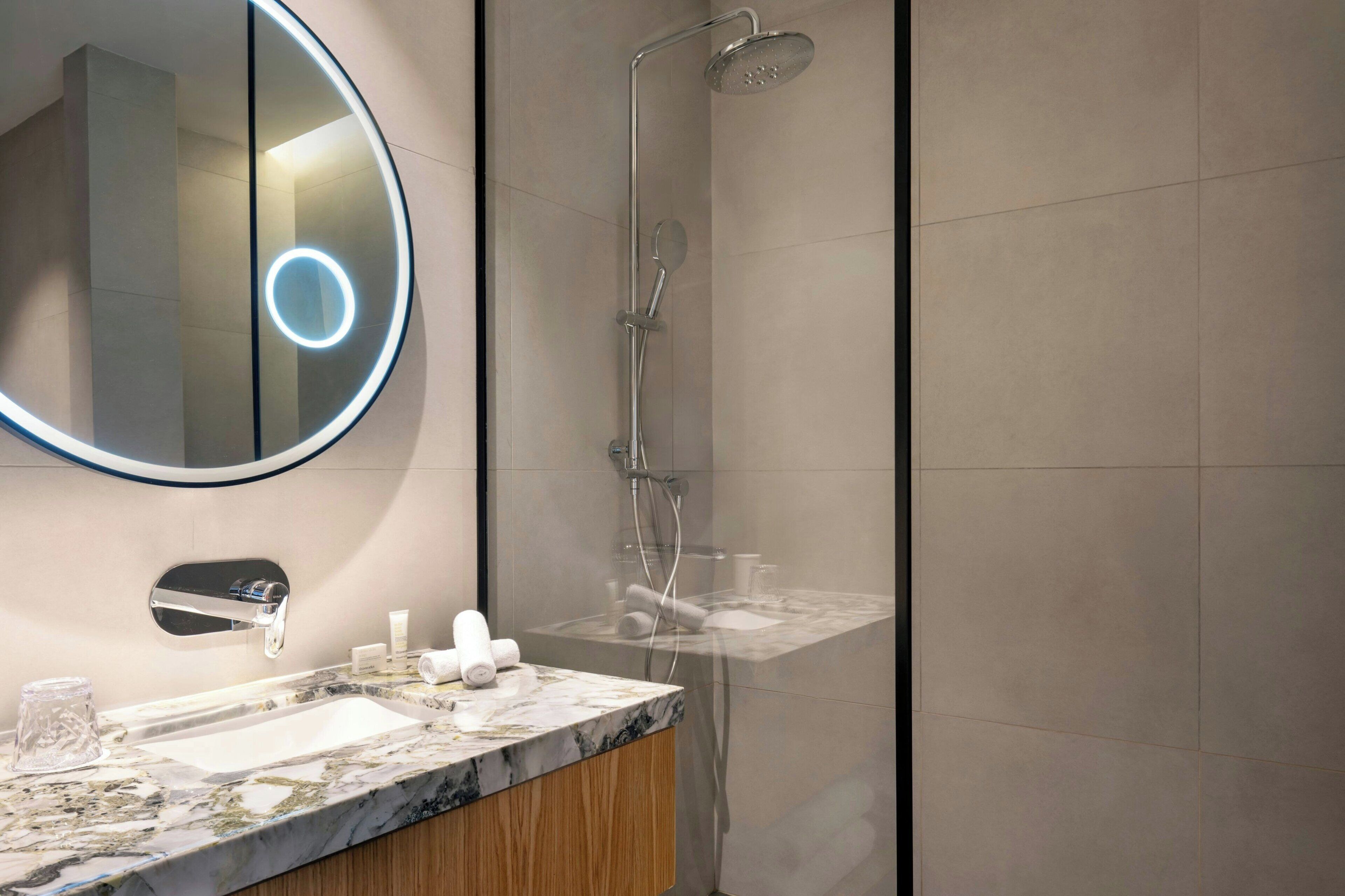A modern bathroom with a marble sink, a round mirror with a light, and a shower.